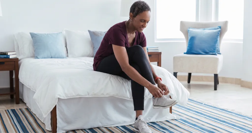 An older woman sits on her bed tying her running shoes, practicing proper shoe fit and foot health every day.